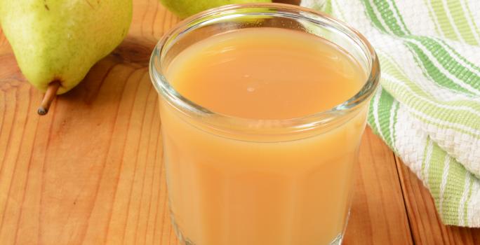 Fotografia em tons de marrom, laranja, branco e verde de uma bancada de madeira marrom vista de cima, contém um copo transparente com suco de pera e maçã. Ao fundo duas peras e ao lado um pano branco e verde