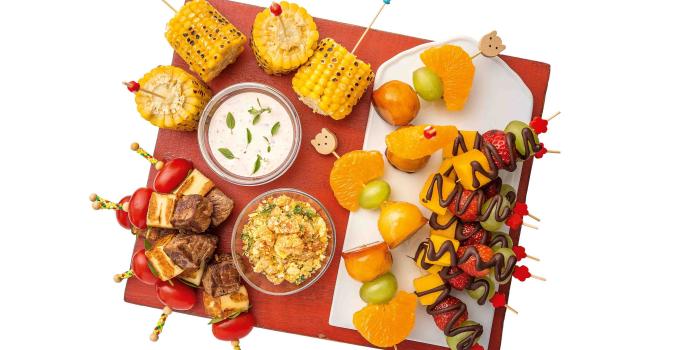 Fotografia em tons de vermelho em uma bancada branca com fundo branco, uma tábua vermelha com vários espetinhos de legumes, carne, frutas com chocolate.