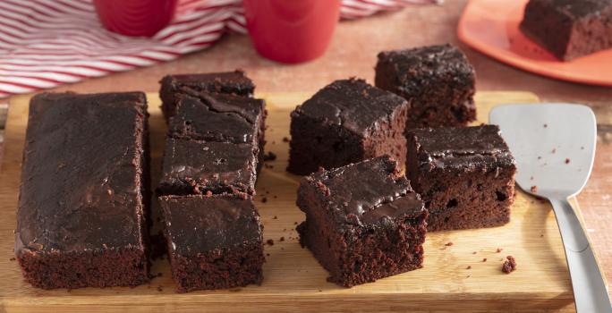 Fotografia em tons de marrom e vermelho de uma bancada de madeira, ao centro uma tábua com fatias de bolo de chocolate. Ao fundo 2 xícaras e um prato vermelho com uma fatia de bolo.