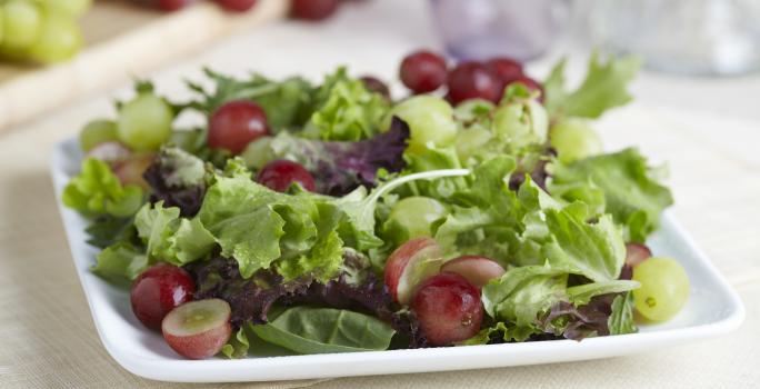 Fotografia de um refratário branco quadrado raso com uma salada de alface, agrião salsão e uva itália com molho de iogurte. O recipiente está apoiado em uma toalha de mesa em tom de bege. Ao fundo tem dois cachos de uva, sendo uvas verdes e uvas roxas.