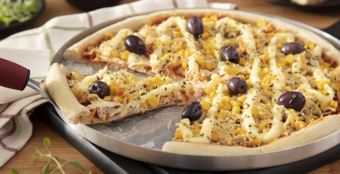 Fotografia em tons amarelo em uma bancada de madeira, uma tábua de alumínio redonda com a pizza de frango com catupiry em cima dela. Ao lado, potinhos com milho, azeitonas e um prato com uma fatia da pizza.