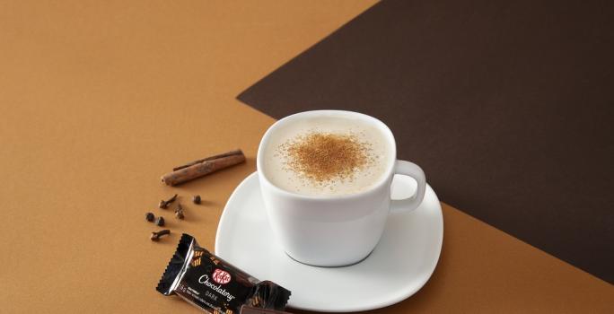 Foto da receita de cappuccino chai cremoso servida em uma xícara branca com um kit kat dark ao lado em fundo marrom claro e escuro