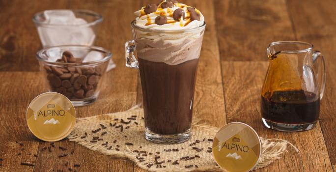 Fotografia em tons de marrom em uma bancada de madeira com um copo de vidro grande e a bebida de chocolate Alpino dentro. Ao fundo, um pote com chantilly, outro com a calda de caramelo e outro com gotas de chocolate e as cápsulas Dolce Gusto Alpino.