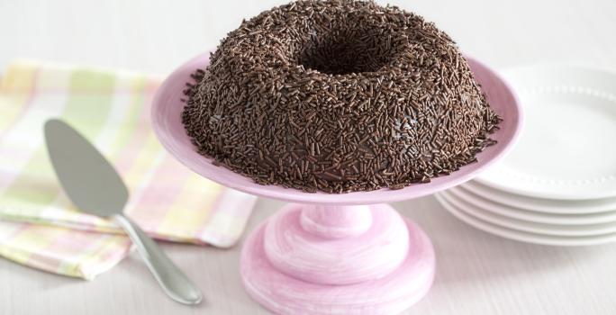 Foto de um brigadeirão de chocolate em cima de um prato de bolo rosa e com pratos brancos atrás