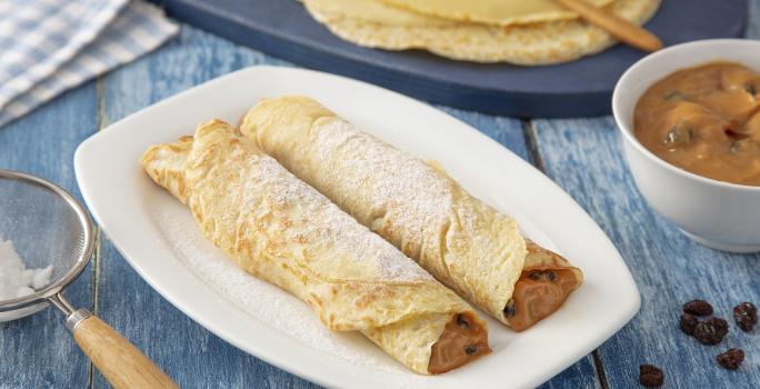 Foto da Receita de Panqueca de Doce de Leite. Observa-se um prato quadrado branco com duas panquecas em cima polvilhadas com açúcar. Do lado esquerdo, tem-se uma peneira com o açúcar e, do direito, as massas de panqueca inteiras.