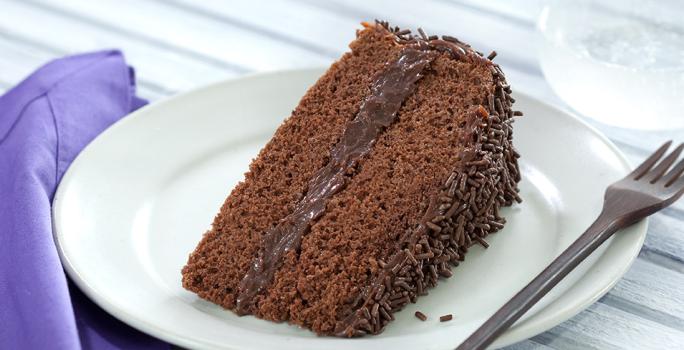 Foto de um prato branco numa mesa de madeira branca com uma fatia de bolo de chocolate dentro e um garfo do lado