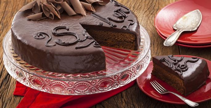 Fotografia em tons de vermelho em uma bancada de madeira com um suporte para bolo de vidro com a torta sacher de chocolate em cima cortada ao meio. Ao lado, pratinhos vermelhos e um deles com uma fatia da torta.