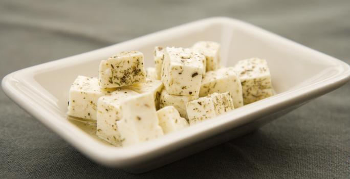 Fotografia de cubos de queijo branco fresco temperados e com azeite dentro de um recipiente pequeno, fundo e quadrado de vidro na cor branco. O aperitivo está sobre uma toalha de mesa preta.