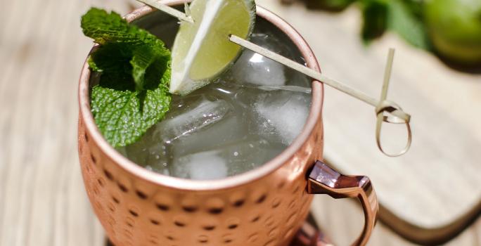 Foto da receita de Moscow Mule Blue, servida em uma caneca de estanho com gelo, sobre uma bancada de madeira decorada com algumas fatias de limão