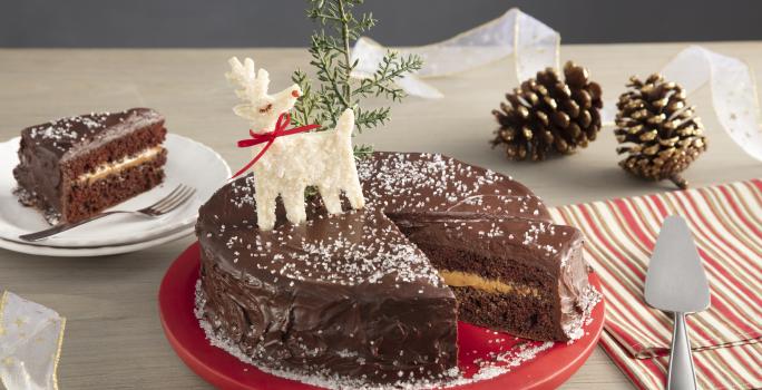 Fotografia em tons de vermelho em uma bancada de madeira clara, um pano bege com listras vermelhas, um prato vermelho redondo com o bolo de chocolate com recheio de doce de leite com decoração de natal. Ao fundo, um prato com uma fatia do bolo.