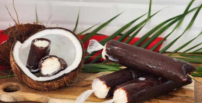 Fotografia de geladinhos em sacos plásticos com cobertura de chocolate e creme de coco branco por dentro. Ao lado cocos decorativos.