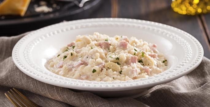 Foto em tons de cinza e branco de uma bancada de madeira escura com uma pano cinza dobrado, um garfo dourado envelhecido e uma prato redondo branco com o risoto dentro. Ao fundo, um prato preto e uma jarrinha de azeite.