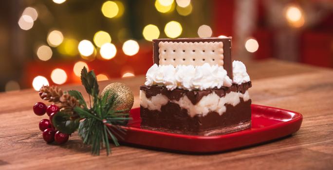 Fotografia de um bolo com três camadas com chantilly e um biscoito no topo. O bolo está sobre um prato vermelho.