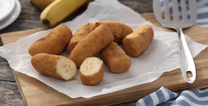 Fotografia em tons de amarelo e azul em uma bancada de madeira clara, um pano azul com listras brancas, uma tábua de madeira com várias bananas à milanesa em cima dela. Ao lado, uma concha de fritura e ao fundo um cacho de bananas.