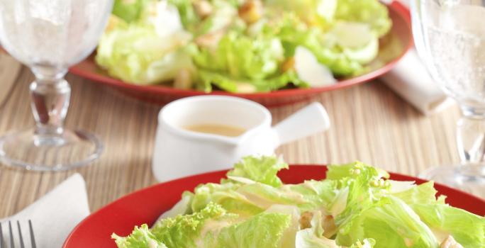 Fotografia em tons de vermelho e verde em uma bancada de madeira clara com um prato vermelho ao centro e a salada caesar com croutons em cima dele.