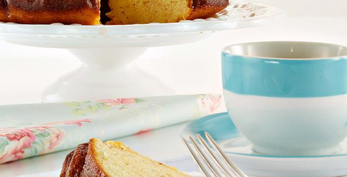 Fotografia em tons de azul em uma mesa branca com uma toalha branca, um prato de sobremesa com uma fatia de bolo de maracujá. Ao fundo, um suporte para bolo com o bolo inteiro e ao lado uma xícara com listra azul e chá dentro dela.