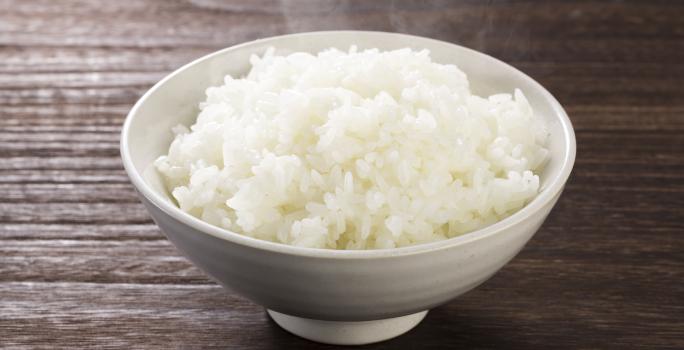 Fotografia em tons de marrom e branco de uma bancada de madeira vista de cima, um recipiente branco redondo contém arroz branco dentro.