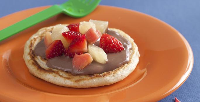 fotografia em tons de laranja e azul de uma bancada vista de frente, contém um prato redondo e laranja com uma panqueca redonda com cobertura de chocolate e frutas picadas por cima.