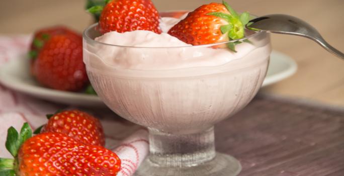 Fotografia de um creme de frutas vermelhas com iogurte, sobre o gelado, dois morangos inteiros e uma colher apoiada no recipiente de vidro em que está o creme. Ao lado, dois morangos, e ao fundo, mais morangos em cima de um prato branco pequeno.
