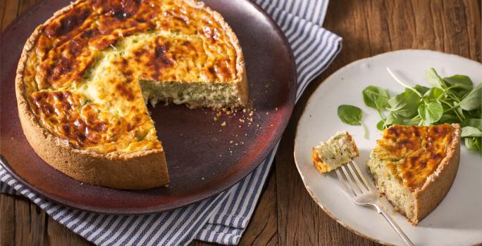 Fotografia em tons de marrom e branco de uma bancada de madeira com um paninho branco e azul, sobre ele um prato redondo vinho com um quiche. Ao lado um prato branco redondo com uma fatia de quiche.