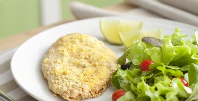 Fotografia em tons de verde em uma bancada de madeira com um pano branco com listras beges e um prato oval branco com o filé de frango empanado e salada verde dentro dele.