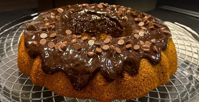 Foto da receita de Bolo de cenoura e especiarias sem glúten. Observa-se um bolo com furo no meio fofinho com uma calda brilhante de chocolate decorado com gotas.