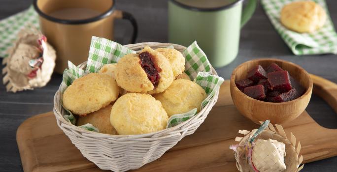 Foto em tons de amarelo e marrom da receita de pão de queijo romeu e julieta servida em diversas porções em uma cestinha sobre uma tábua de madeira com decoração de festa junina e um bowl com goiabada. Ao fundo há duas canecas