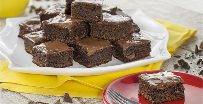 Fotografia em tons de chocolate, branco e amarelo, com prato branco com pedaços de bolo de chocolate sobre guardanapo amarelo, prato vermelho com porção de bolo e um garfo, no alto pote amarelo com chocolate, sobre bancada clara com pedaços de chocolate.