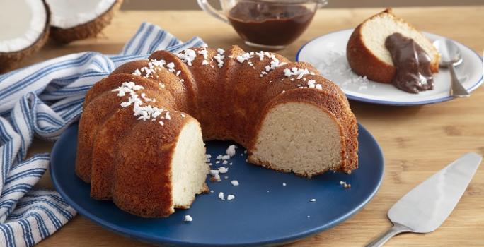 Foto da receita de bolo de leite de coco cremoso caseiro servida em uma porção com uma fatia cortada sobre um prato de porcelana azul escuro com a fatia sobre um prato de sobremesa branco com uma cobertura de chocolate e ao lado um paninho listrado
