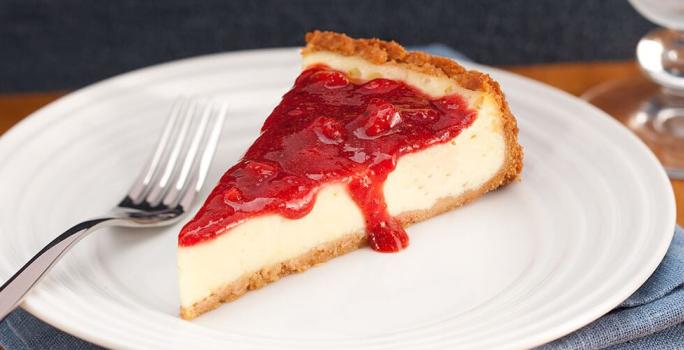 Fotografia em tons de branco e vermelho, no centro na foto está uma fatia da torta coberta com geleia de morango, ao lado está um garfo e embaixo do prato um guardanapo azul.