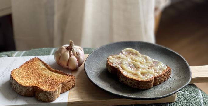 Foto de um prato escuro redondo com uma fatia de pão com a manteiga de alho por cima. Ao lado esquerdo dele há outra fatia de pão torrada e um dente de alho inteiro ao fundo.