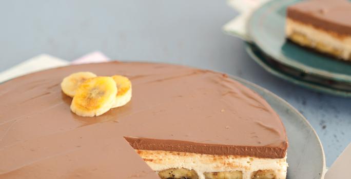 Fotografia em tons azul em uma mesa de madeira azul, com um pano branco com rosa florido, um prato azul raso redondo com a torta mousse de doce de leite e banana.