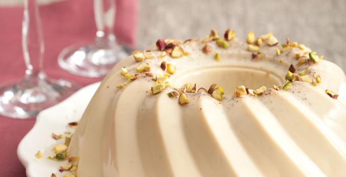 fotografia em tons de cinza e bege vista de frente, contém um prato redondo branco com um Flan de pistache e ao lado duas taças transparentes,  abaixo um pano cor vinho