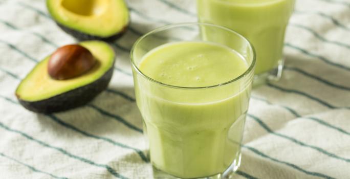 Fotografia de dois copos de vidro com vitamina de abacate. Ao fundo, ao lado do segundo copo, tem um abacate cortado ao meio, sobre uma toalha de mesa branca com listras verdes.