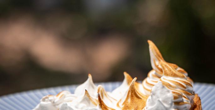 Fotografia de uma fatia de torta de cupuaçu feita com creme de leite, e por cima tem um merengue maçaricado. A fatia está em um prato de vidro branco com bordas cor na cor azul. Ao fundo da foto tem algumas árvores.