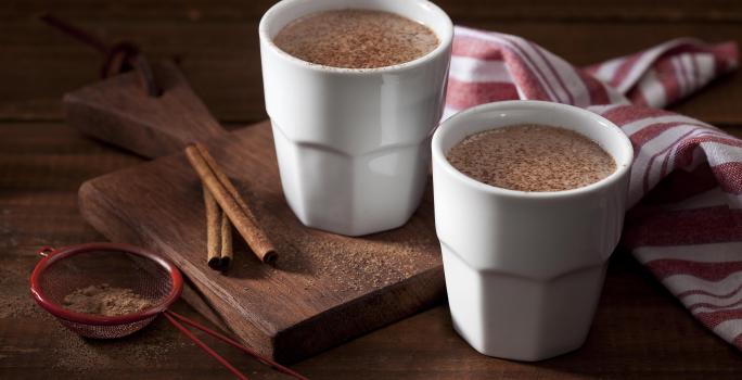 Foto dois copos brancos com chocolate quente dentro em cima de uma tábua de madeira com canela em pau e em pó decorando a mesa