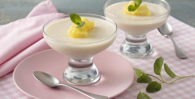 Fotografia em tons de rosa e amarelo de uma bancada, ao centro um paninho quadriculado rosa e branco, um prato rosa com uma taça com o mousse e pedacinhos de abacaxi e folha de hortelã. Ao lado uma taça igual e folhas de hortelã.