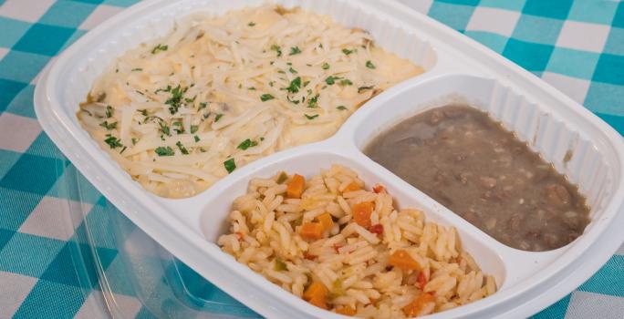 Foto aproximada de uma marmita branca de plástico com três divisões, uma com o frango com creme de milho, outra com arroz e outra com feijão. Ela está sobre um pano azul e branco quadriculado