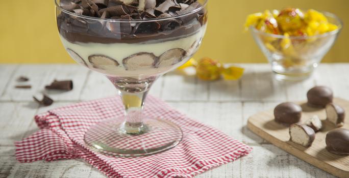 Fotografia em tons de amarelo e chocolate, ao centro taça com creme, bombons cortados ao meio e raspas de chocolate sobre guardanapo xadrez branco e vermelho, ao lado tábua com bombons, ao fundo potinho com bombons, tudo em bancada de madeira em pátina.