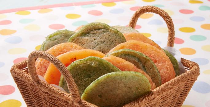 Fotografia em tons de verde e laranja em uma bancada de madeira, uma toalha branca com bolinhas coloridas, uma cesta de vime marrom clara com pães de queijo achatados nas cores verde e amarelo.