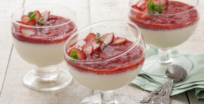 Fotografia em tons de branco, verde e vermelho de uma bancada de madeira branca com um paninho verde com colheres, sobre ele taças de vidro com mousse e purê de morangos.