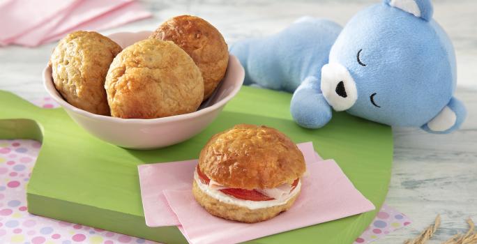 Foto da receita de Pão de Leite. Observa-se um potinho rosa com três pãezinhos e, ao lado direito, um pão com recheio de tomate, requeijão e peito de peru, sobre uma tábua verde.