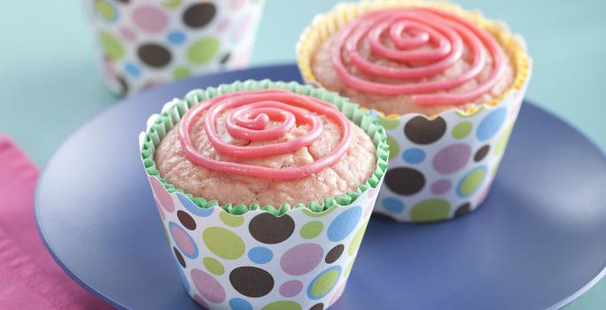 Fotografia em tons de azul e rosa em uma mesa de madeira azul, um pano rosa, um prato azul redondo com dois cupcakes de morango com aveia em forminhas coloridas.