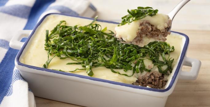 Fotografia em tons de azul em uma bancada e madeira, um pano xadrez azul e ao centro um recipiente refratário branco com detalhes em azul e o escondidinho de carne e batata e couve por cima para decorar.