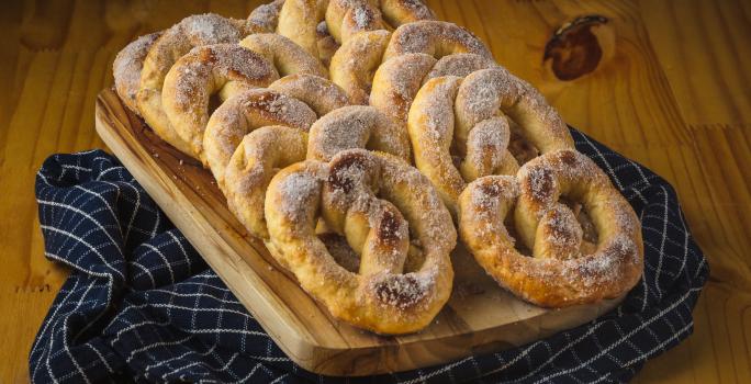 Foto aproximada de uma bancada de madeira sobre a qual está um pano azul quadriculado e nele há uma tábua de madeira com alguns pretzels decorados com açúcar e canela