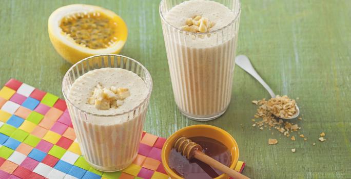 dois copos transparentes com a vitamina dentro ao lado um potinho redondo na cor amarelo com mel dentro e ao fundo uma fatia de maracujá