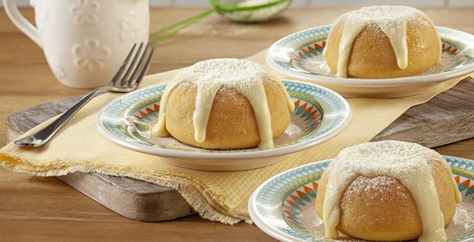 Fotografia em tons de bege com três porções de bolinhos com cobertura em pratinhos coloridos, sobre guardanapo amarelo e tábua de cozinha com um garfo ao lado, pote de cobertura e peneirinha verde ao fundo em bancada de madeira.