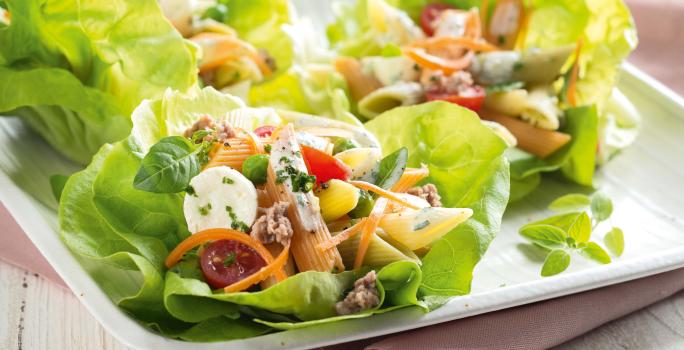 Fotografia em tons de branco e verde, ao centro salada de folhas verdes com macarrão, legumes e mozarela, sobre guardanapo rosa e bancada de madeira em tons de branco.