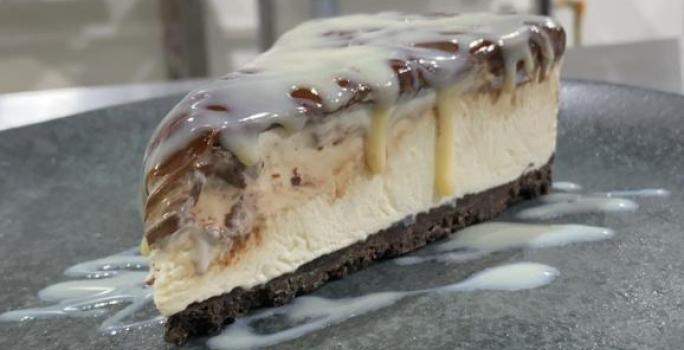 Foto da receita de CHEESECAKE MOÇA GANACHE. Observa-se uma fatia da cheesecake em um prato de cerâmica preto, com Leite MOÇA escorrendo pelo recheio.