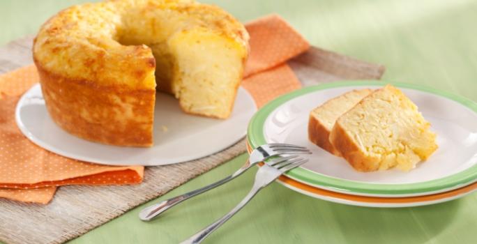 Fotografia em tons de laranja e verde em uma mesa de madeira verde com um pano laranja de bolinhas branca. Ao centro, um prato raso branco com o bolo de macaxeira cortado. Ao lado, um prato branco com borda verde e duas fatias do bolo.